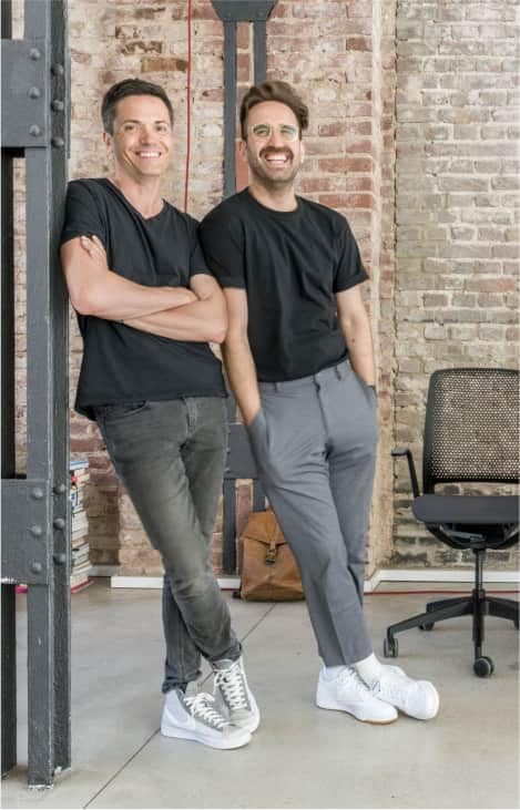 2 männliche Mitarbeiter nebeneinander, einer lehnt an Pfeiler, anderer an seinem Kollegen