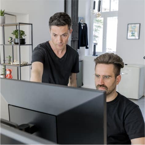 2 männliche Mitarbeiter, einer steht und zeigt auf Screen, anderer Mann sitzt und schaut auf Screen