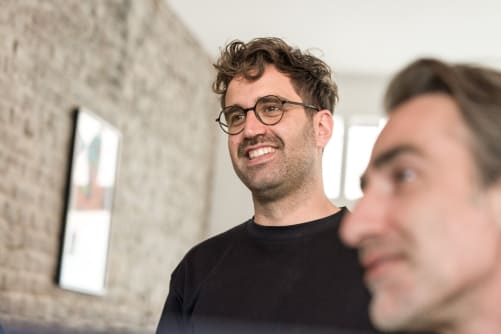 männlicher Mitarbeiter Deni unscharf in Vordergrund, männlicher Mitarbeiter Thomas scharf und lachend im Hintergrund