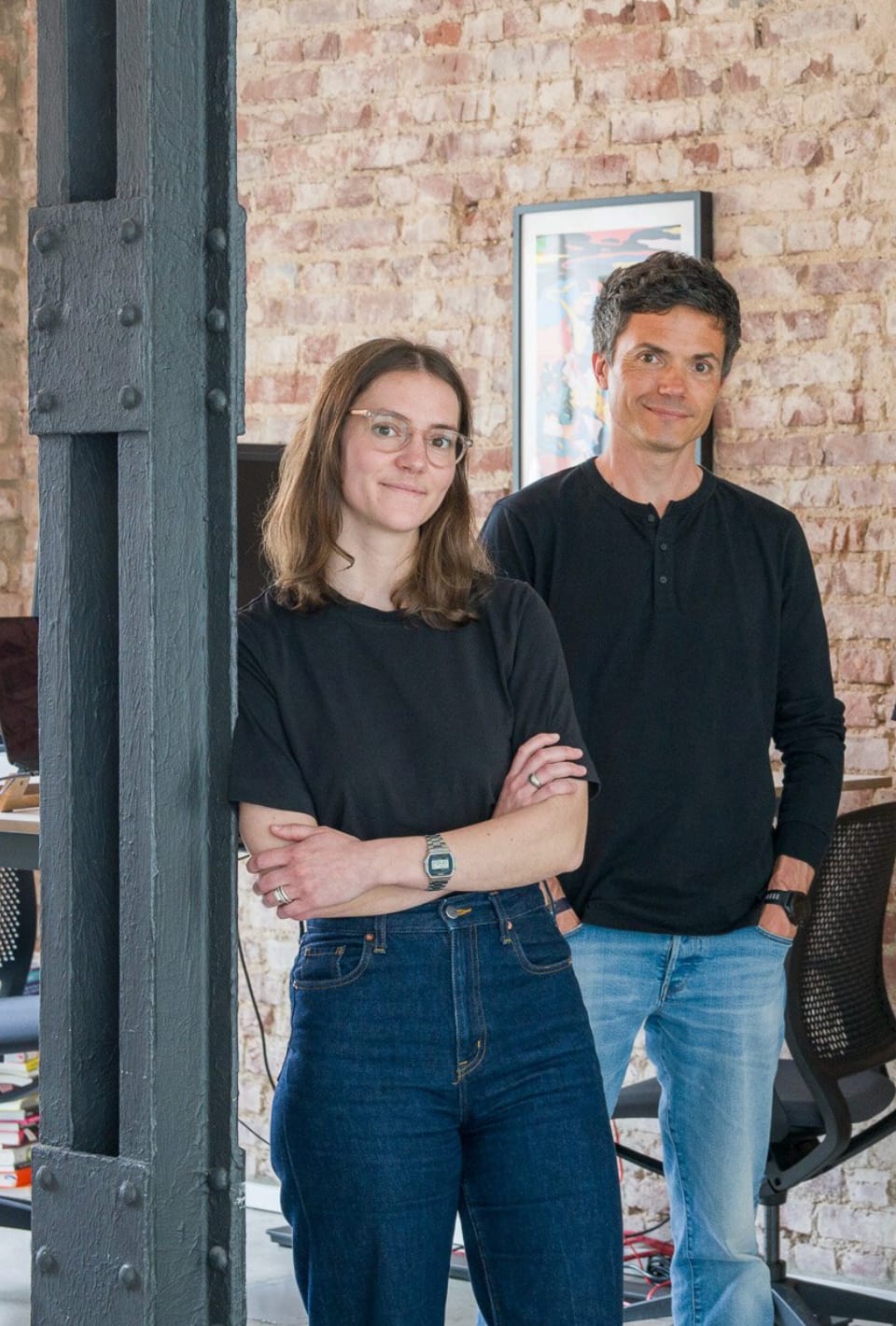 Mitarbeitrin Marie lehnt an Pfeiler dahinter steht Kollege Sebastian grinsend im Hintergrund